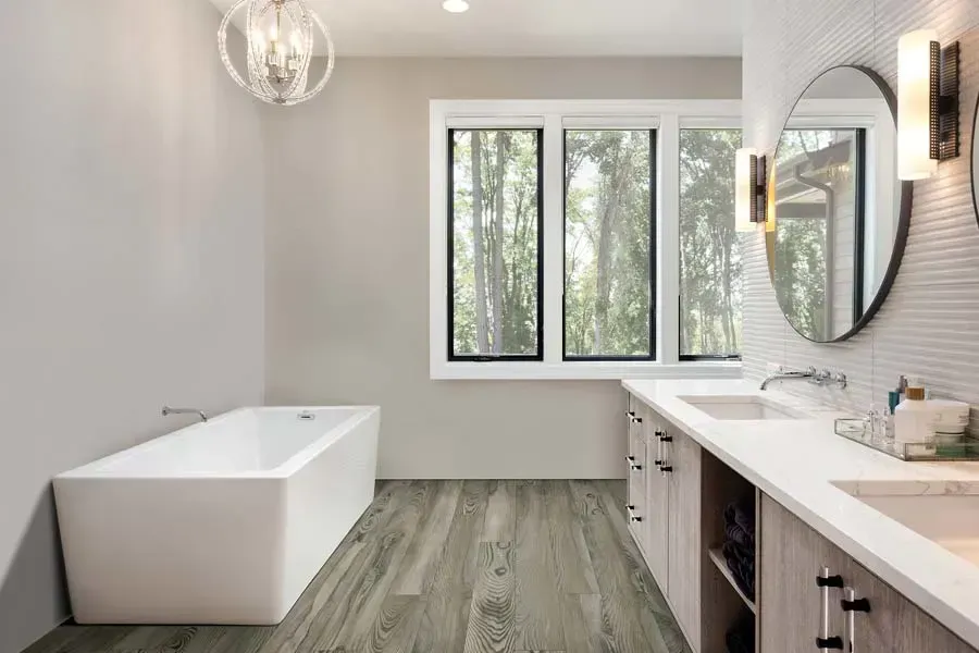 Modern, airy bathroom with a white freestanding tub, light wood vanity, round mirror, and grey wood-look flooring.