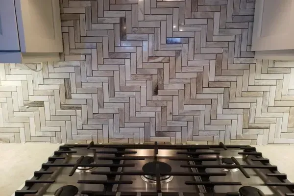 A light gray and beige herringbone-patterned tile backsplash behind a stainless steel gas stovetop in a kitchen.