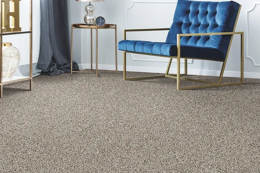 A blue tufted accent chair sits on speckled tan carpet in a modern living room next to a small side table and lamp.