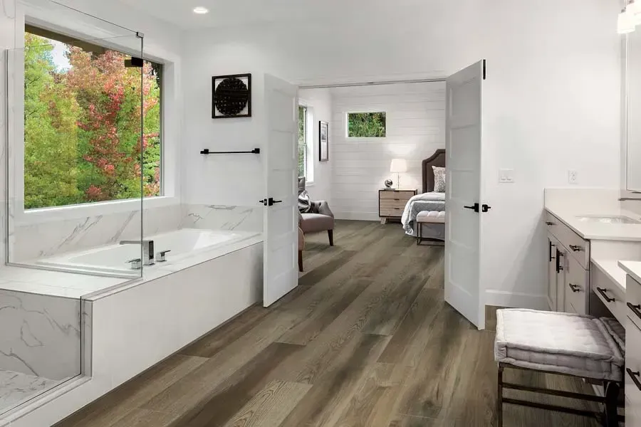 A luxurious bathroom with wood-look flooring, a marble-tiled soaking tub, and open double doors leading to a bedroom.