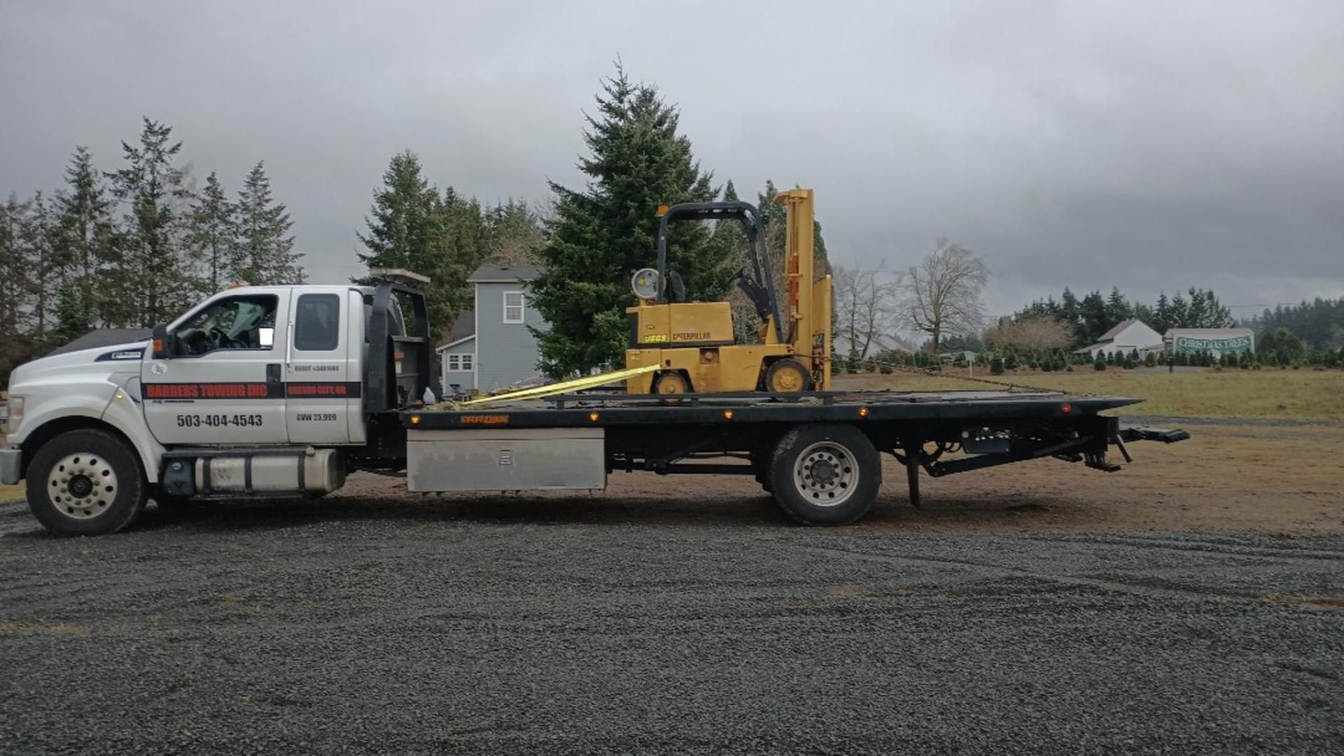 Tow truck with a yellow forklift on its flatbed, outdoors on a gravel lot under an overcast sky.