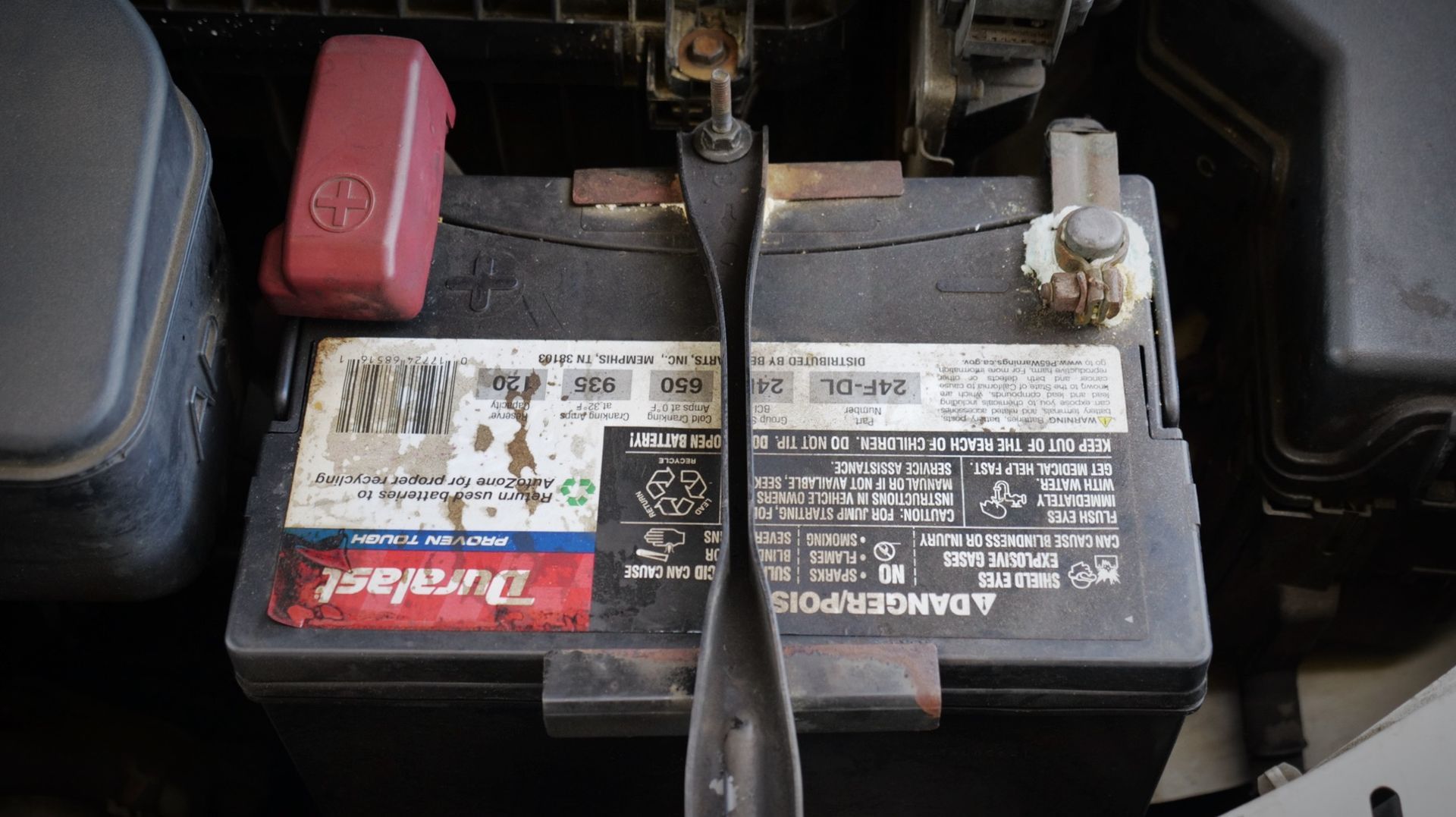 Car battery with hold-down strap. The black battery has a Duracell sticker and red plastic cap.