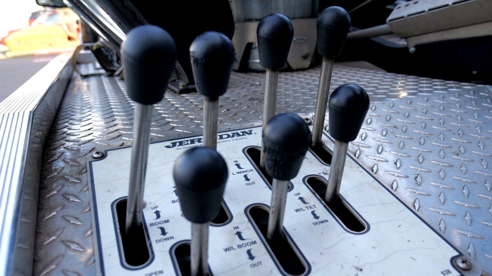 Close-up of a tow truck control panel with seven black-topped levers in a diamond-plated, metal surface.