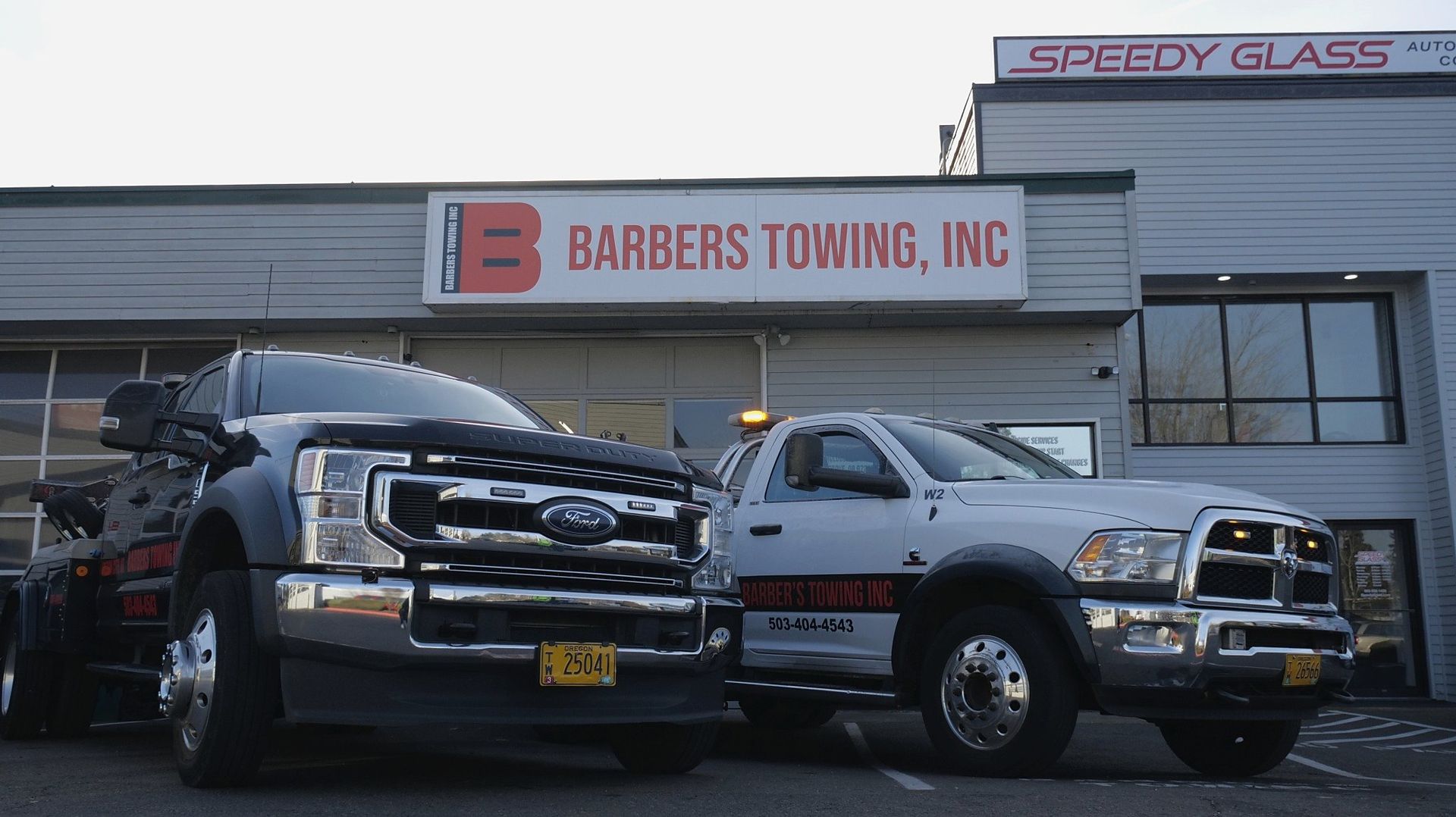 Two tow trucks parked in front of Barber's Towing, Inc. building with Speedy Glass sign in background.