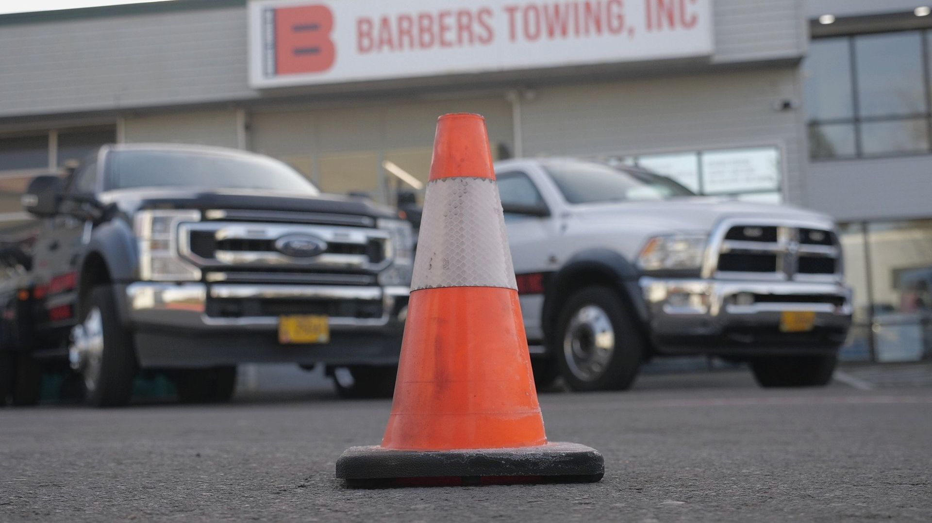 Orange traffic cone on pavement with two tow trucks in the background. Barbers Towing Inc. sign visible.
