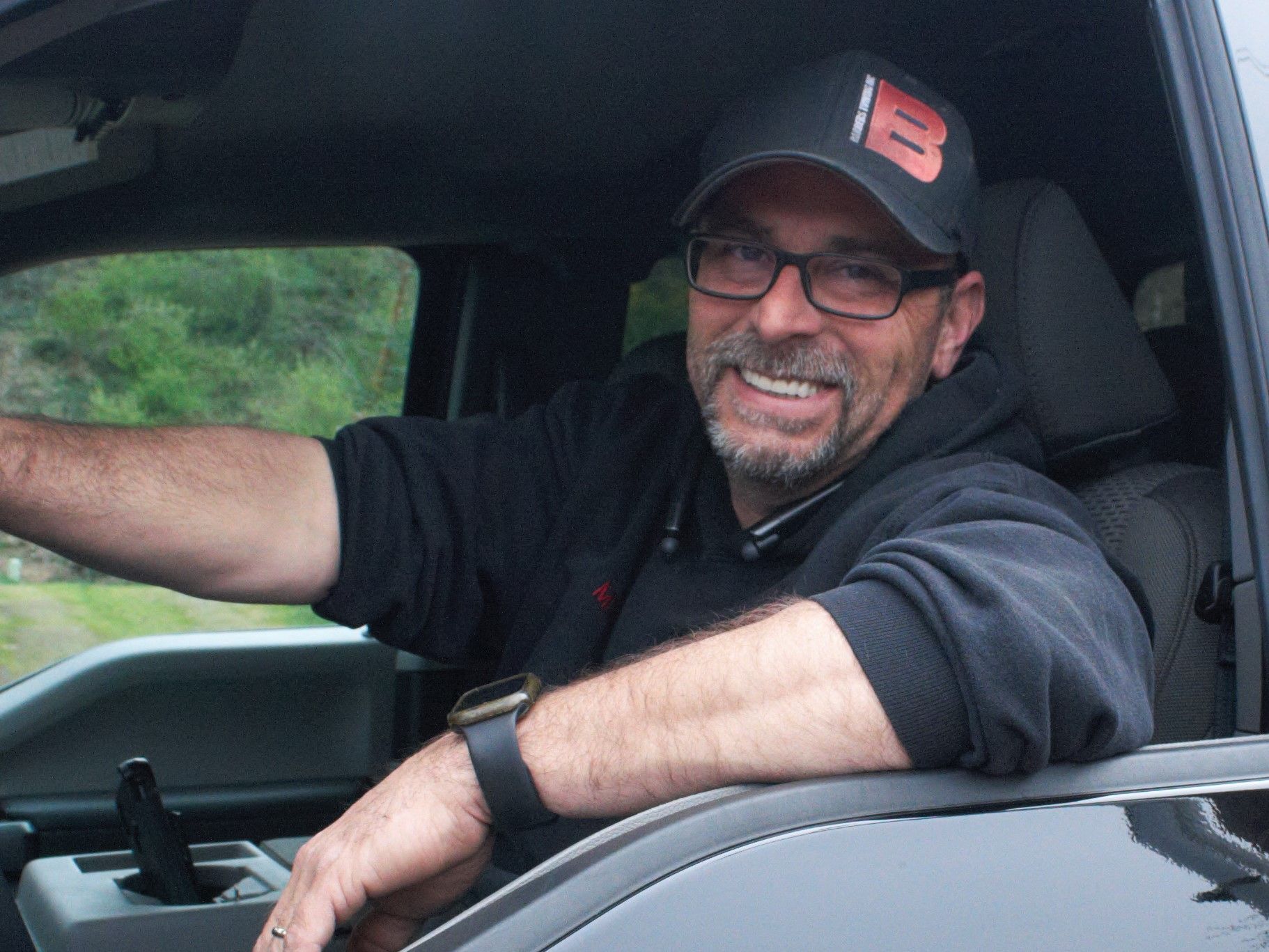 Man in a black hat and glasses smiles from inside a truck.