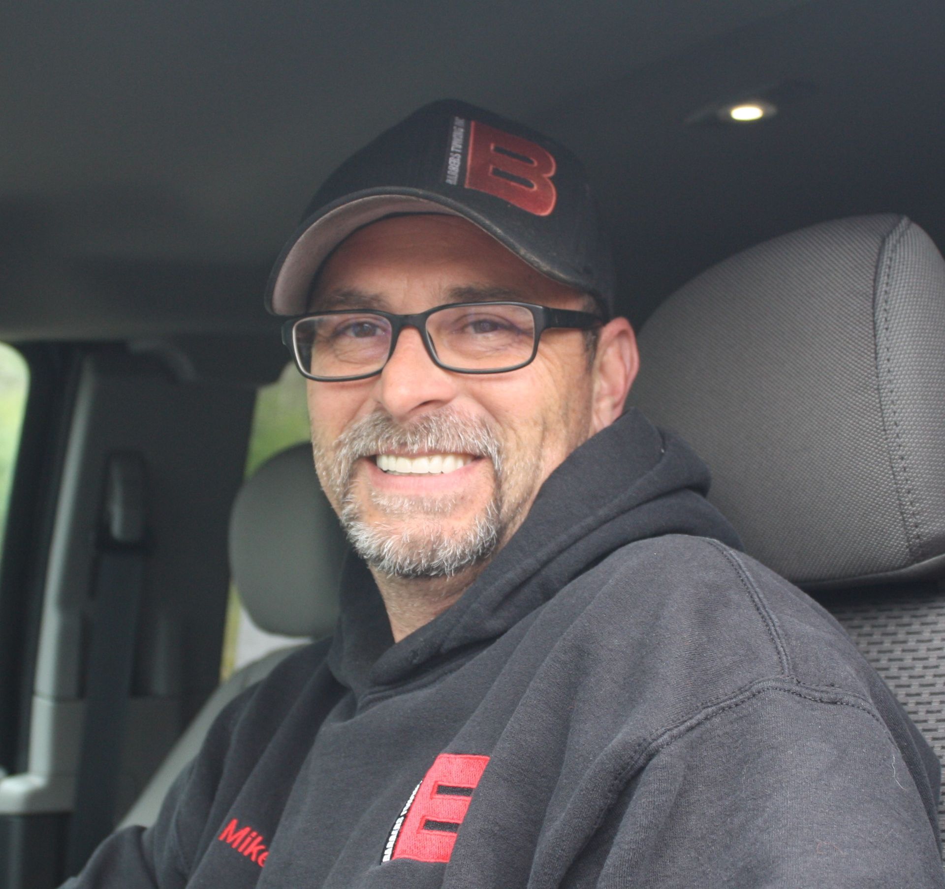 Man in glasses and cap smiles, seated inside a vehicle. He wears a black hoodie with logo.