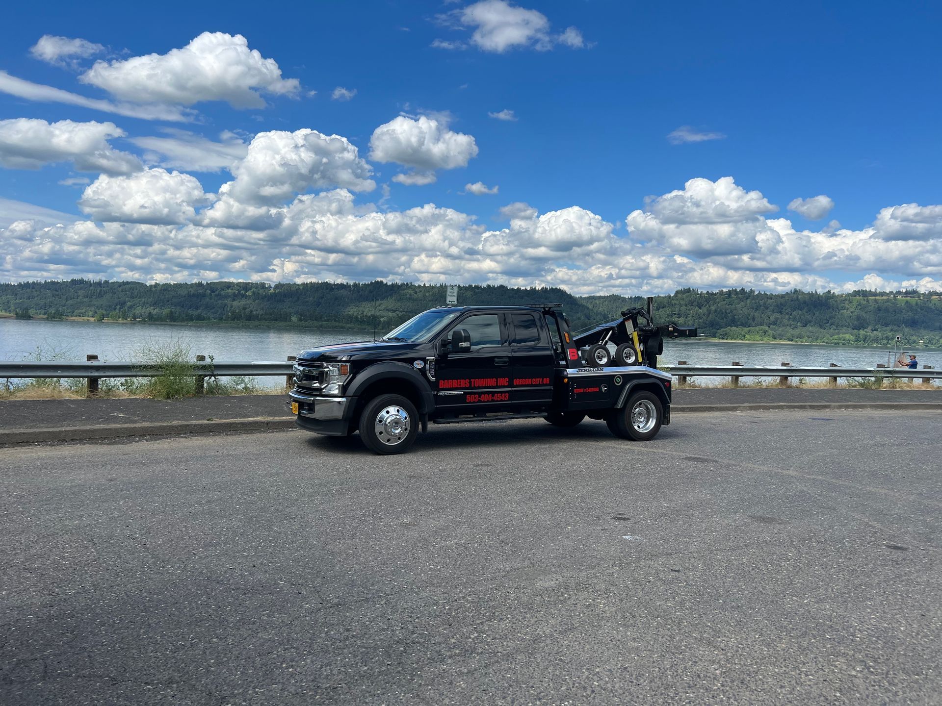 Black tow truck parked on a road by a body of water with a cloudy sky in the background.