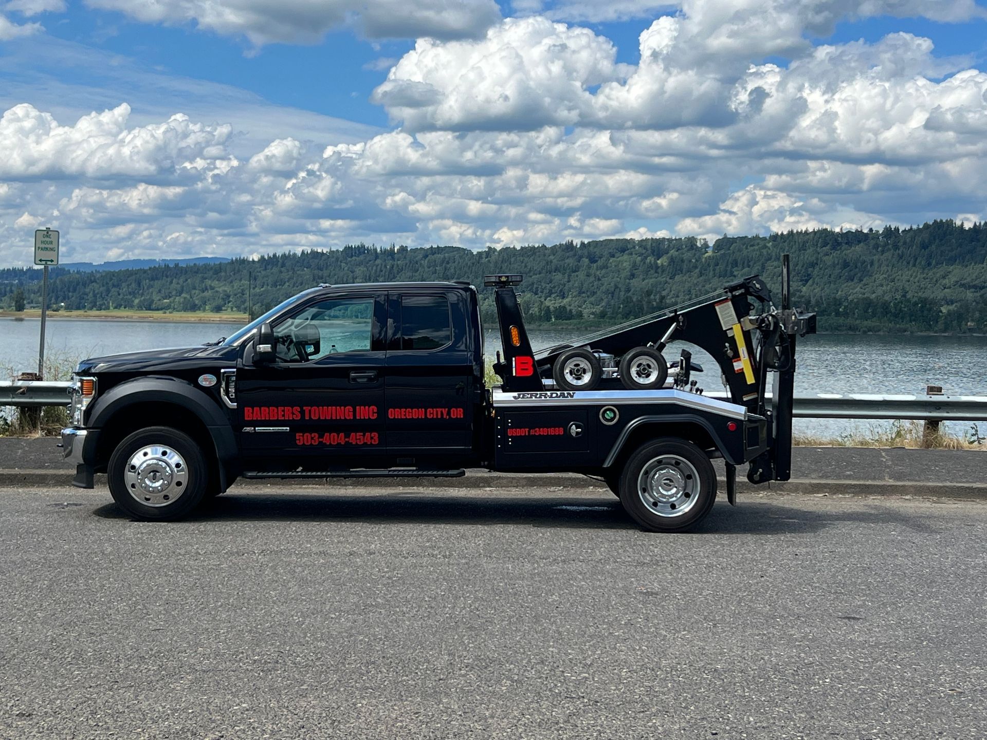 Black tow truck on a road beside a body of water under a cloudy sky.