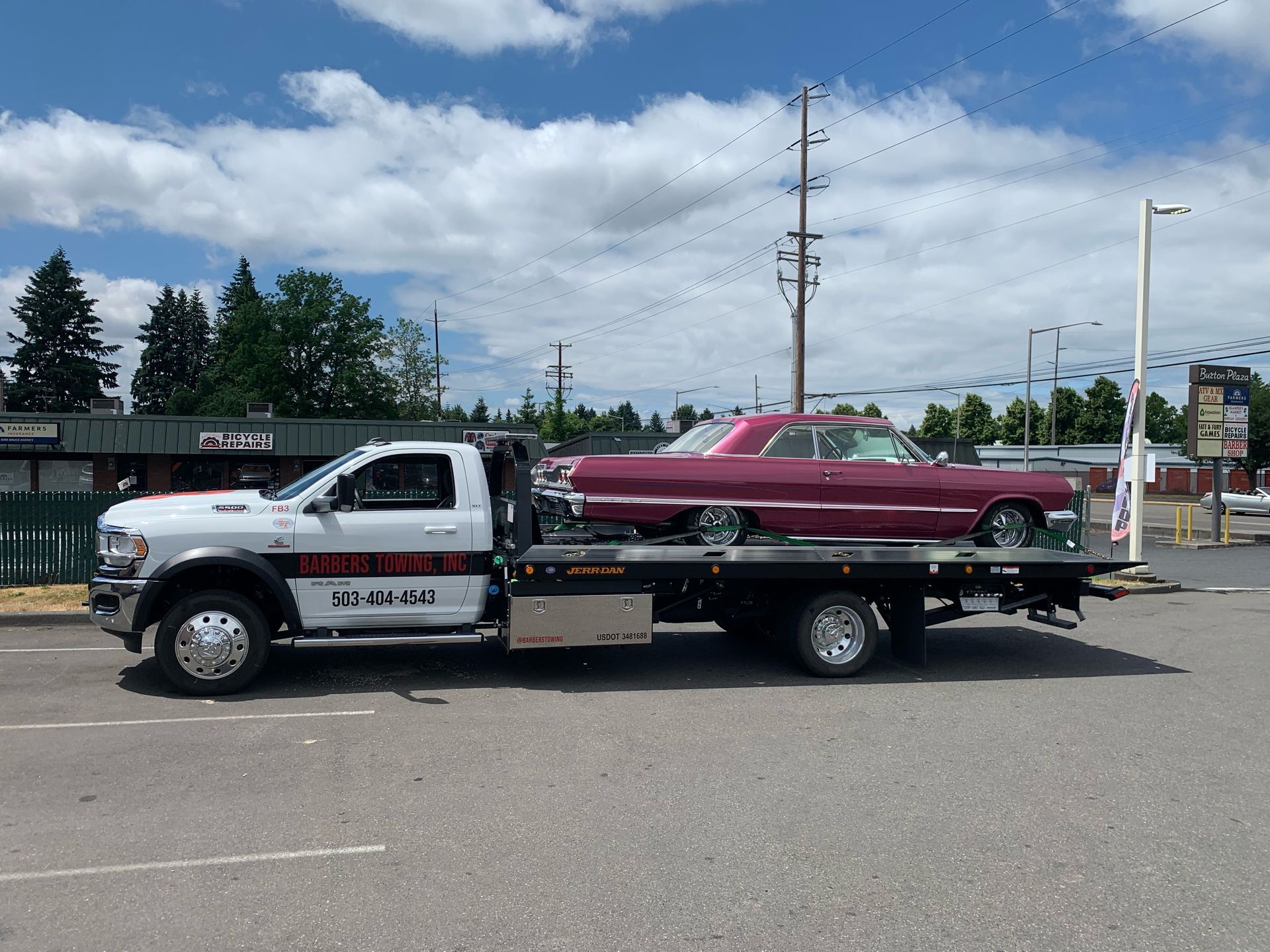 A tow truck carrying a classic maroon car on a flatbed, parked outside a building on a sunny day.