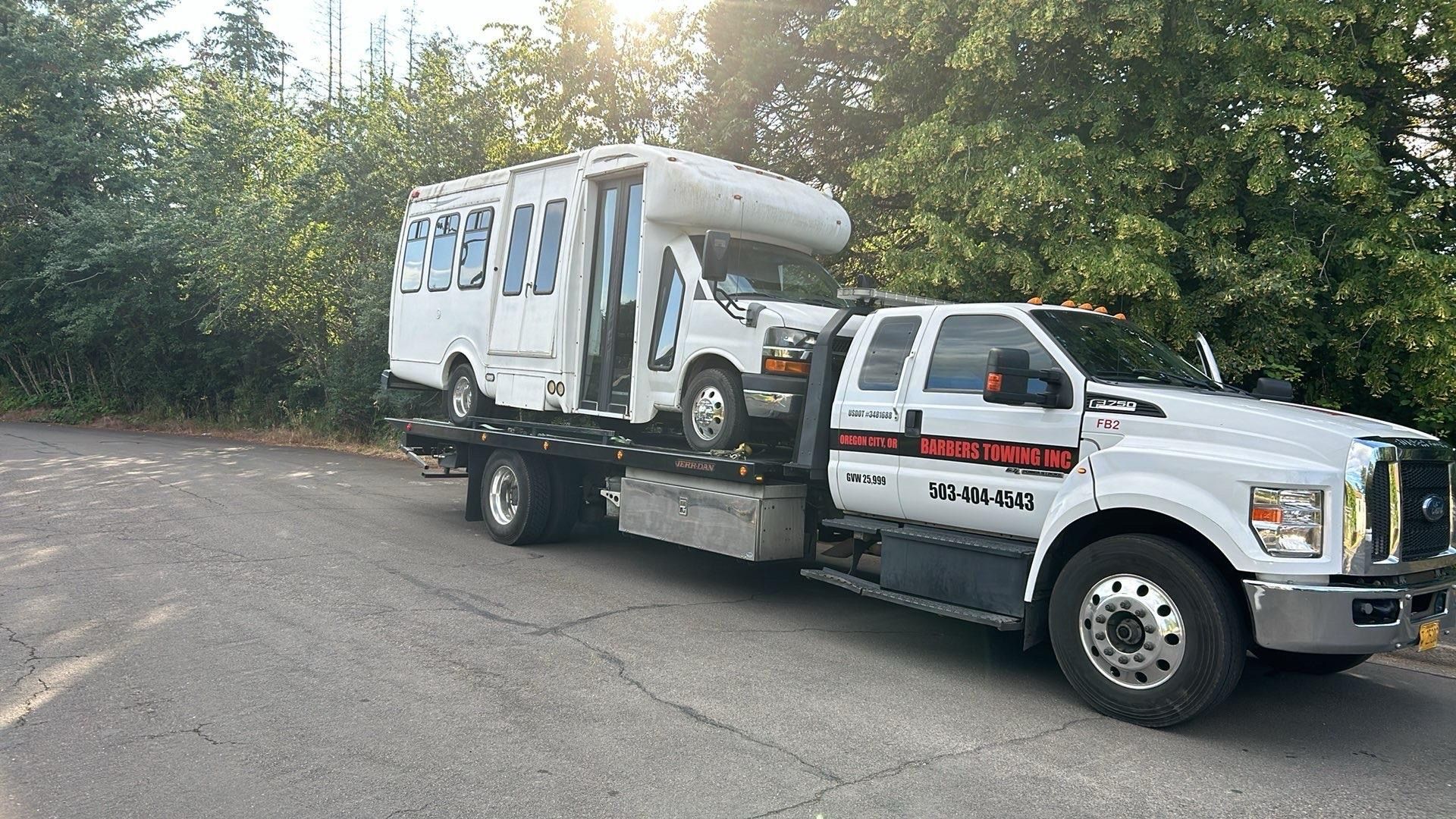 Tow truck carrying a white bus-like RV on a paved road with green trees in the background.