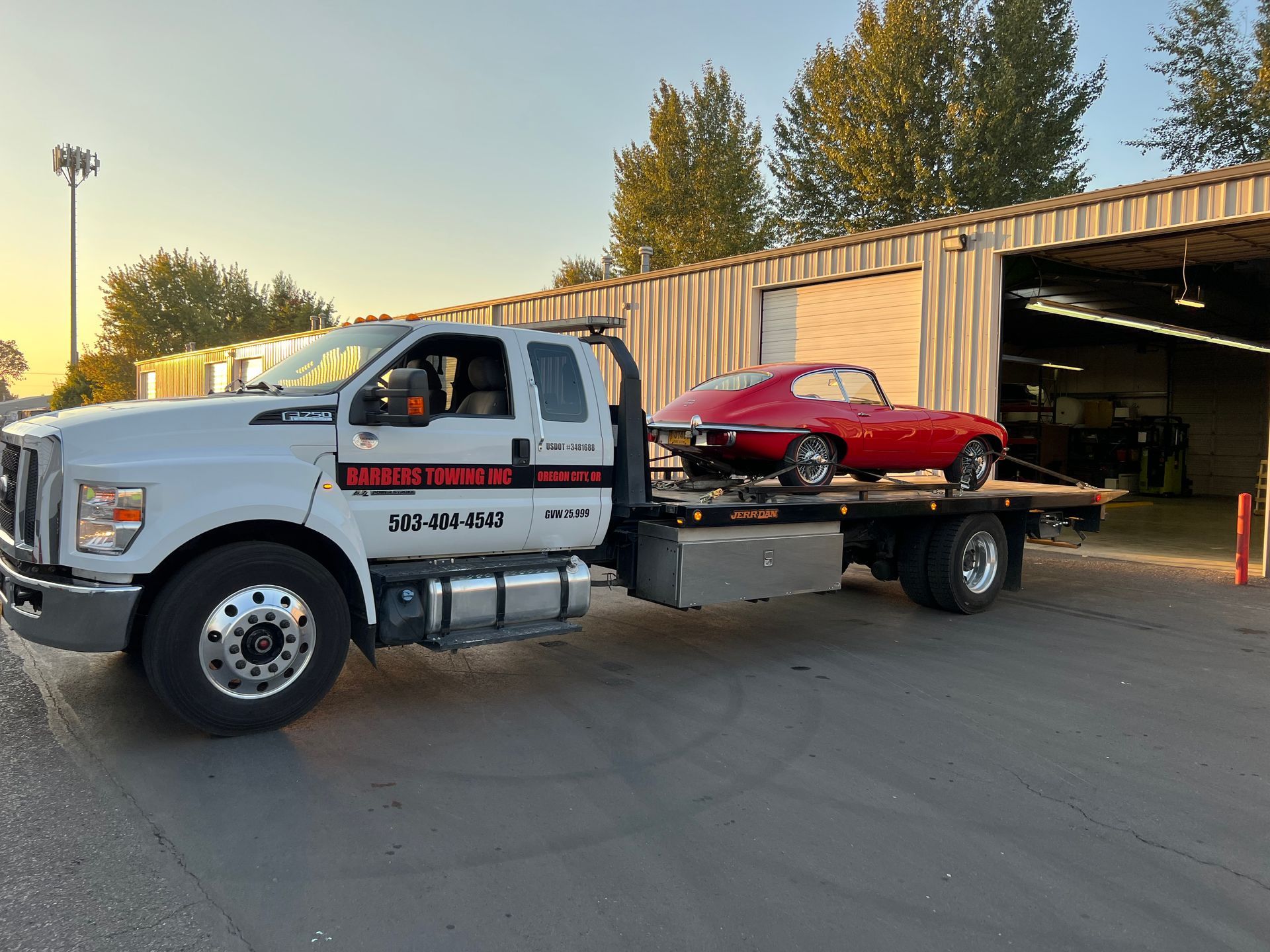 White tow truck carrying a red vintage sports car outside a garage.