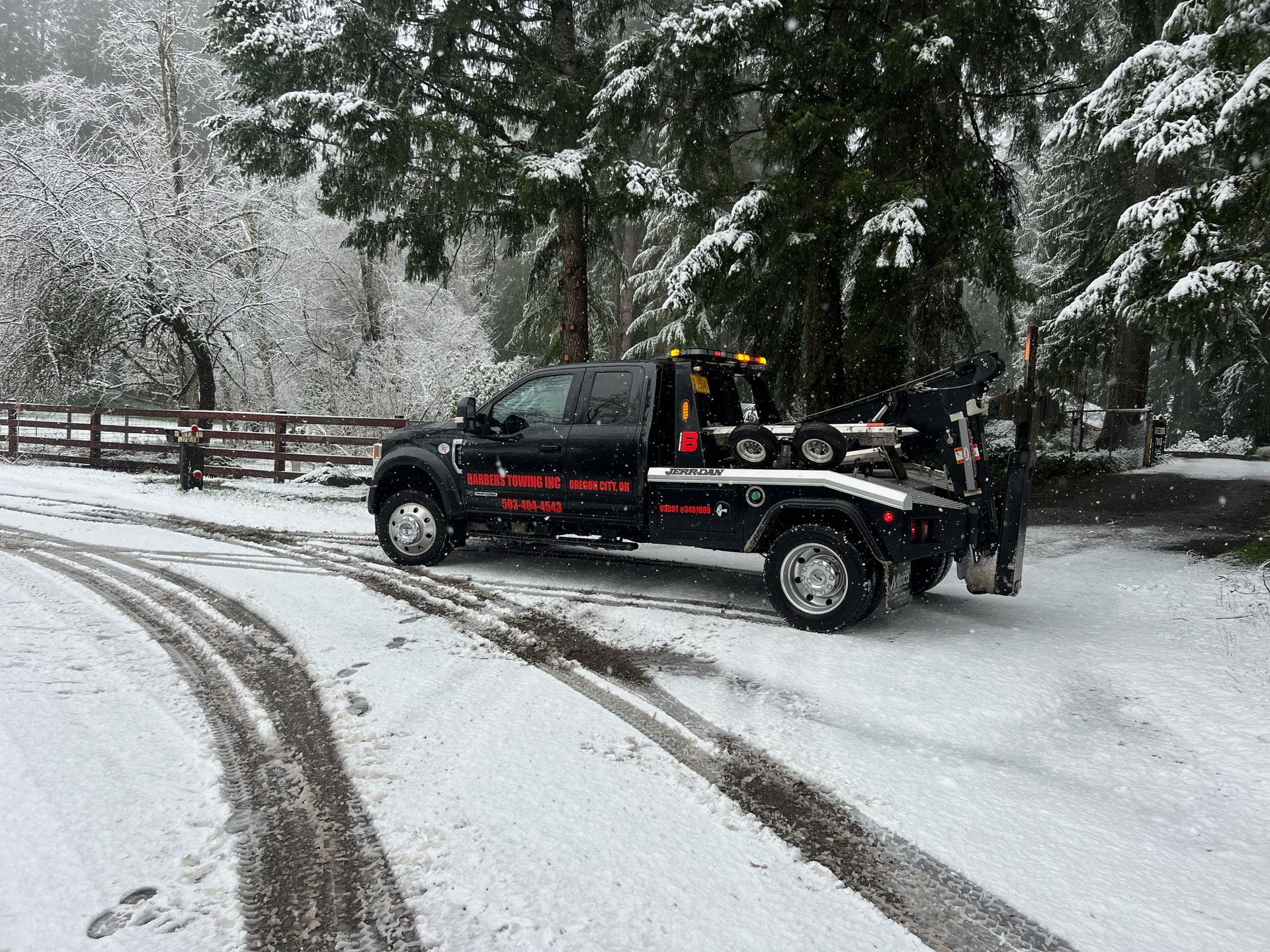Black tow truck on snow-covered road near trees and a fence. Snow falling.
