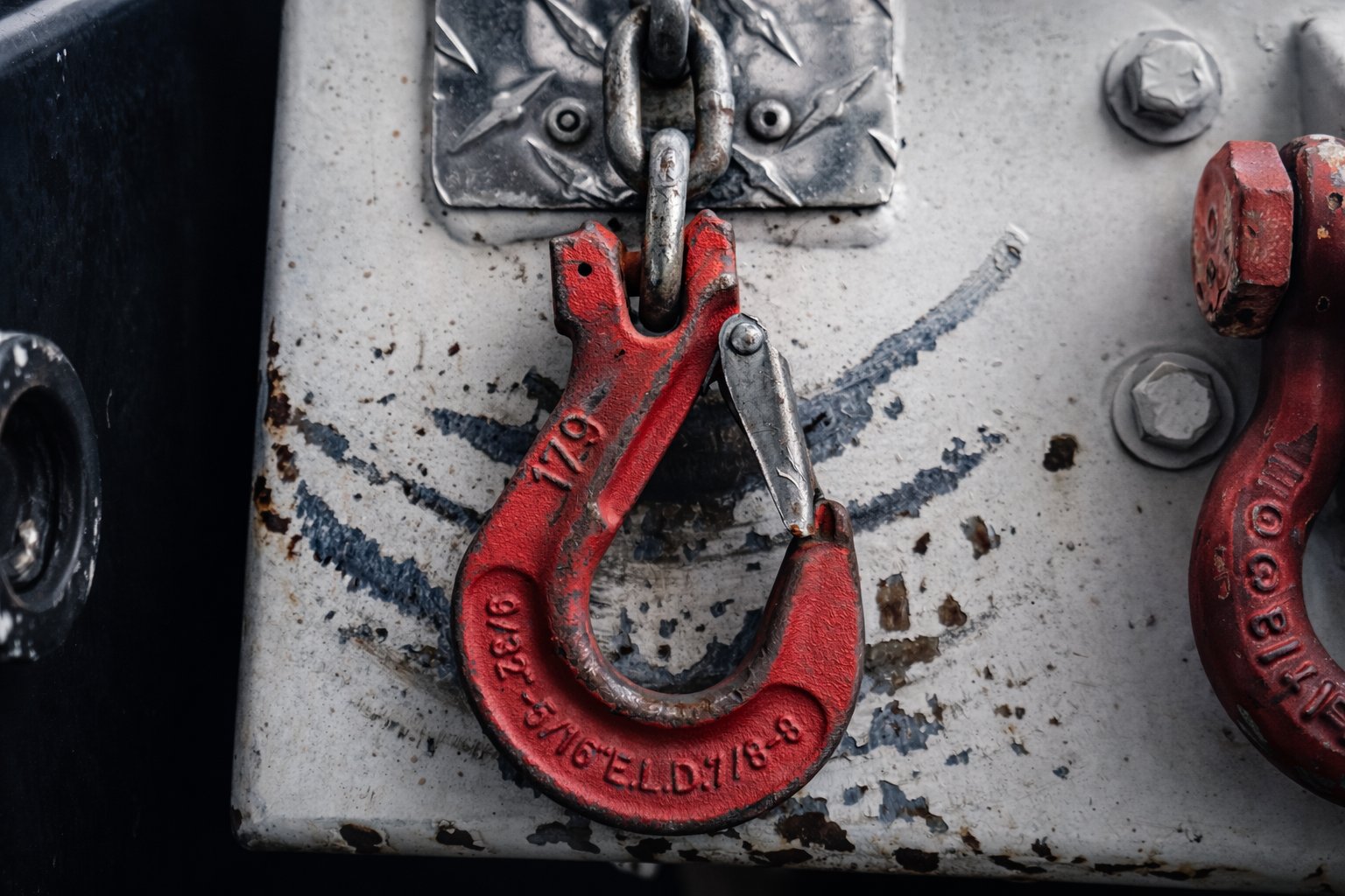 Red metal hook connected to a chain, hanging from a metal surface with bolts.