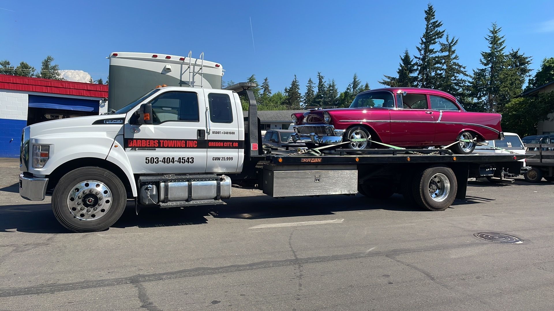 A tow truck carries a pink classic car on its flatbed.  The scene is outdoors on a sunny day.