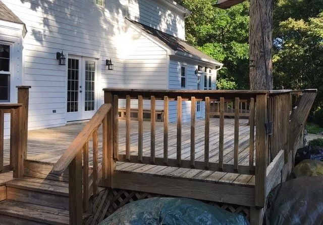 Wooden deck and railing beside a white house with tall windows and trees in the yard