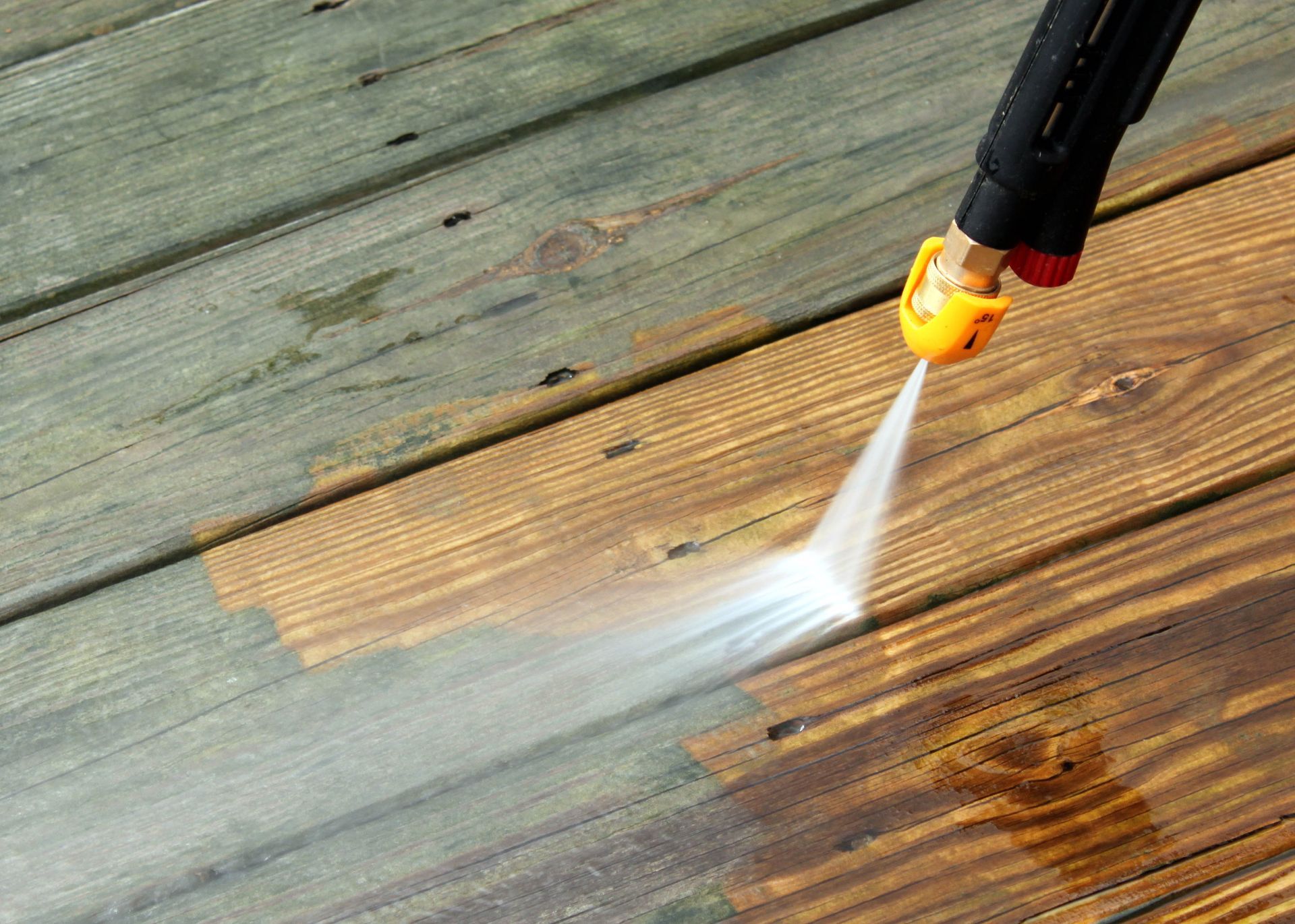Pressure-washing a wooden deck, spraying water across weathered boards with a black and yellow nozzle