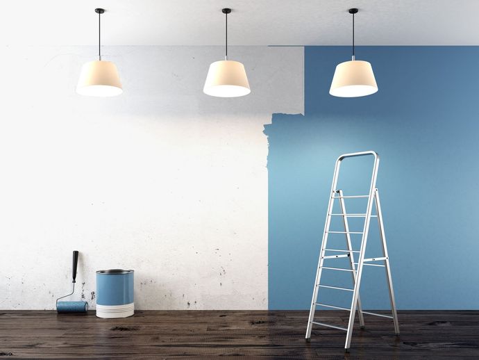 Wall being painted blue with a ladder, paint can, and roller on a wooden floor.