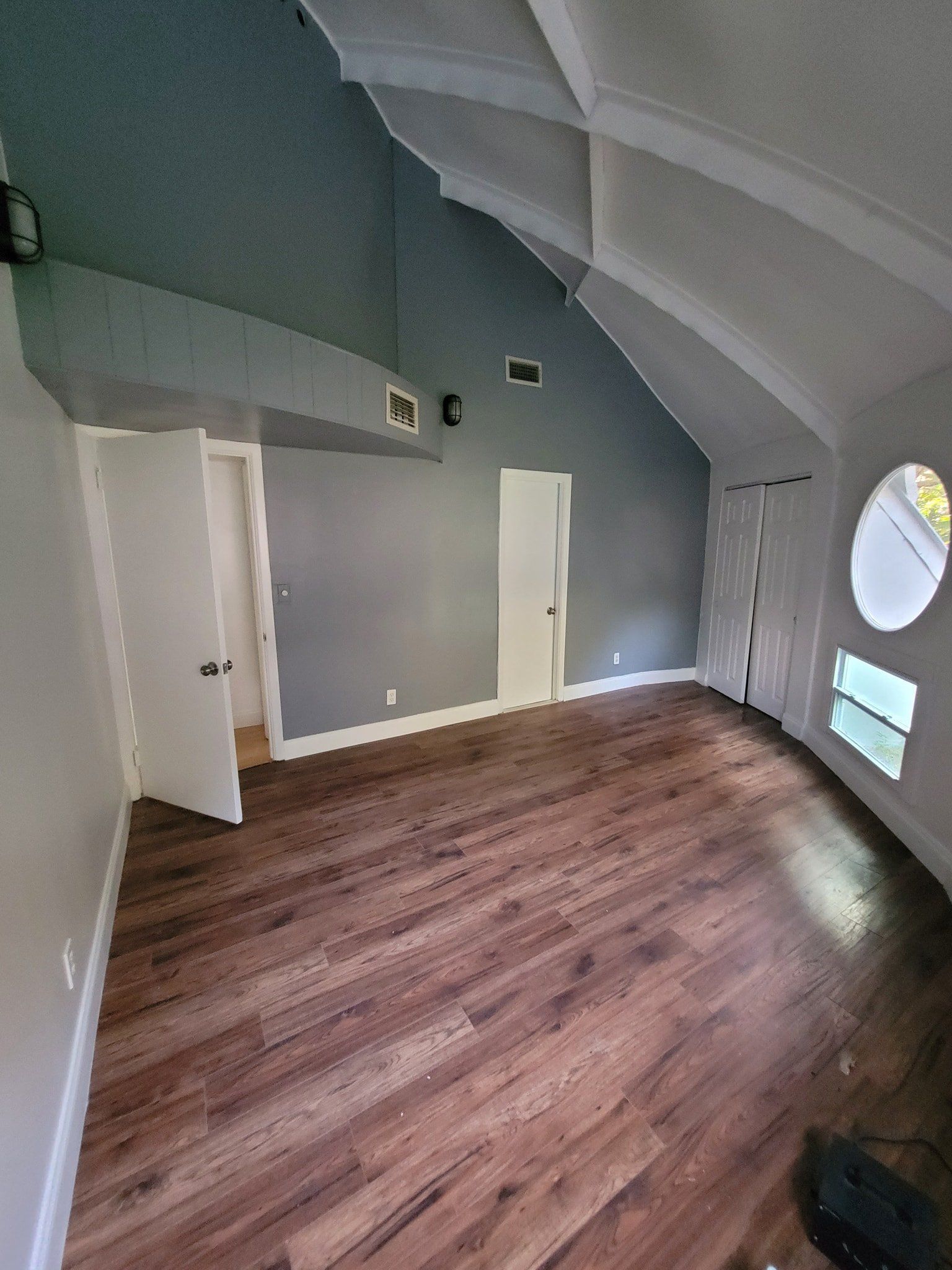 Empty room with hardwood floors, two white doors, and a round window. Grey walls and ceiling.