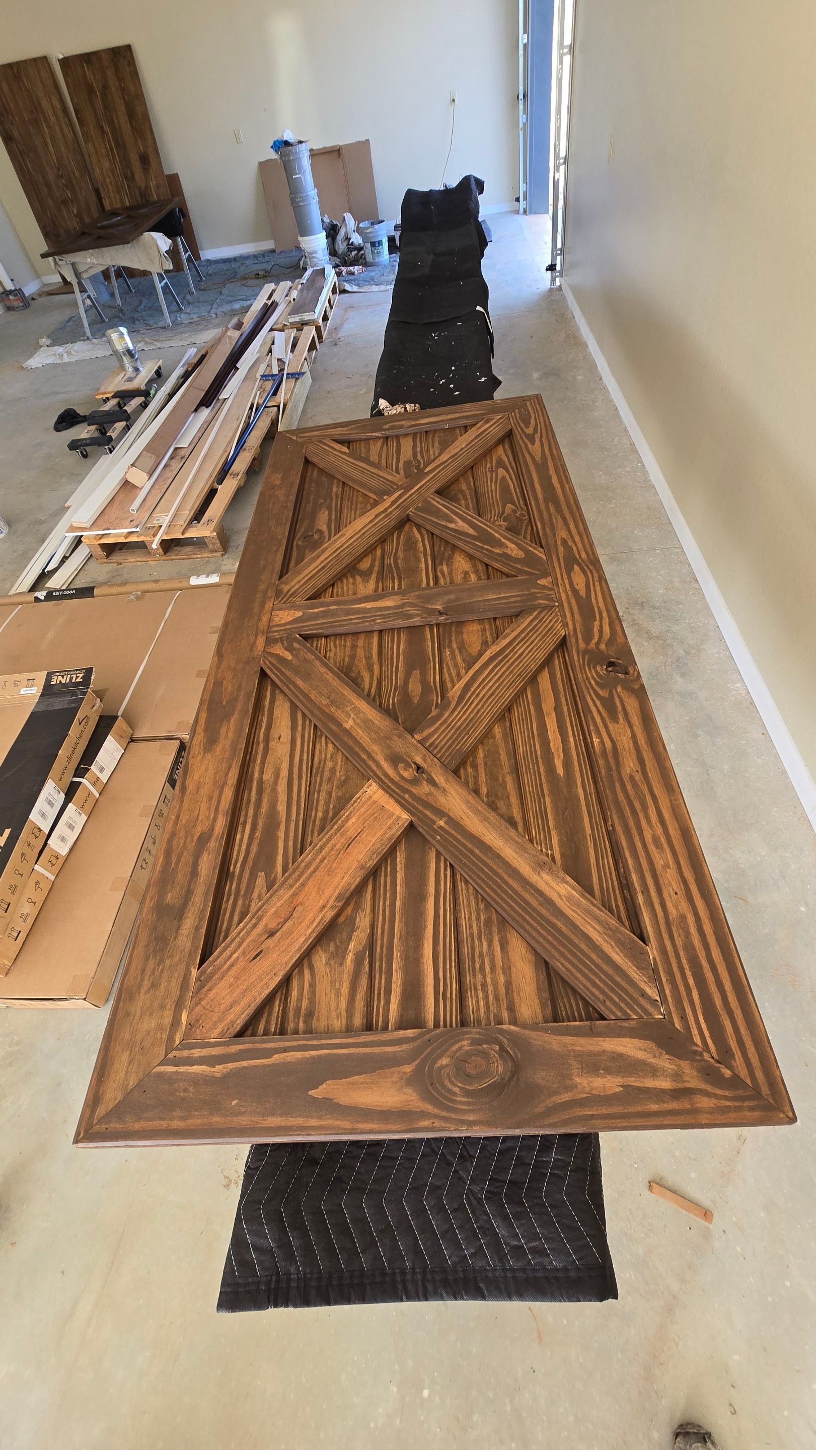 Large brown wooden barn door with an X design, laid out on a black mat in a construction area.