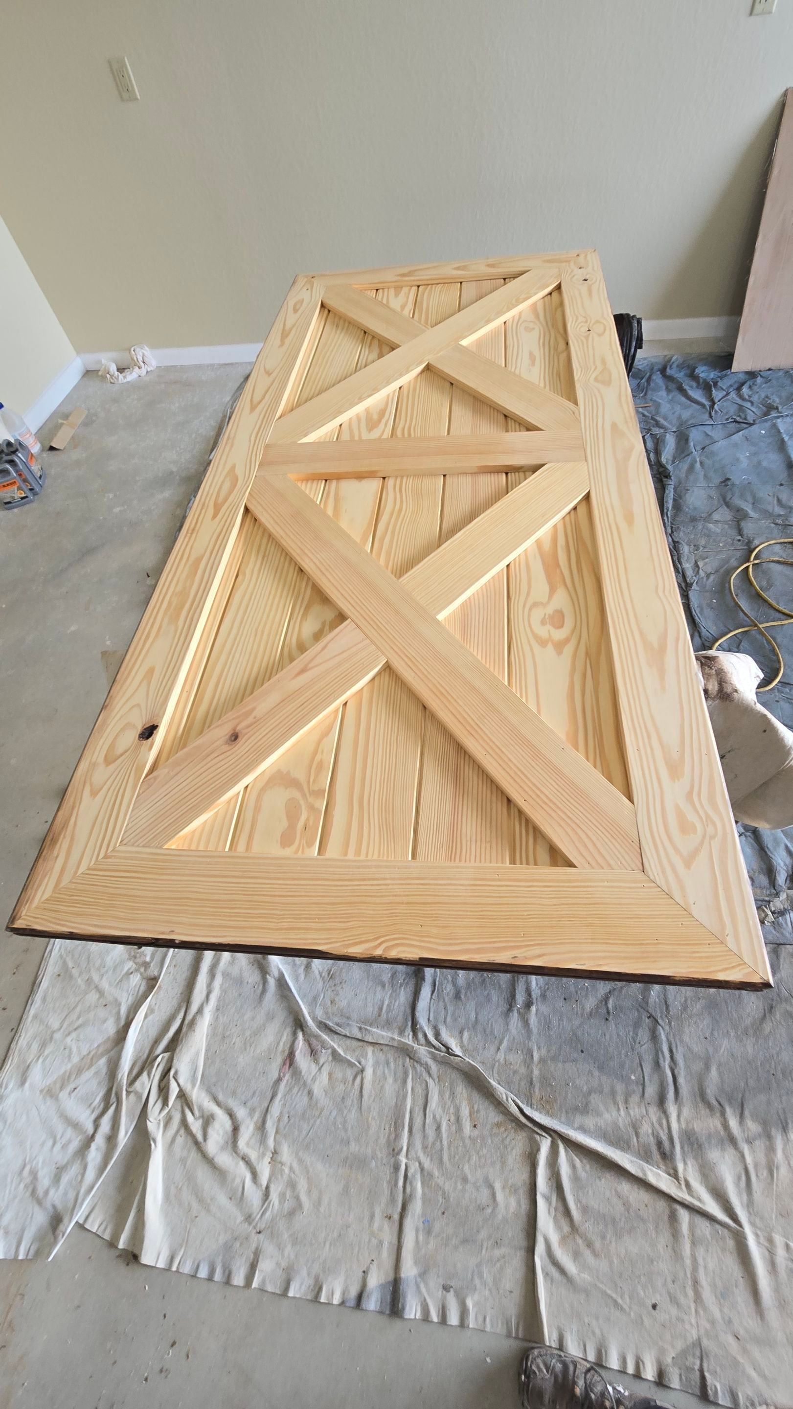 Wooden barn door with an X-shaped design, light-colored, lying on a paint-splattered tarp.
