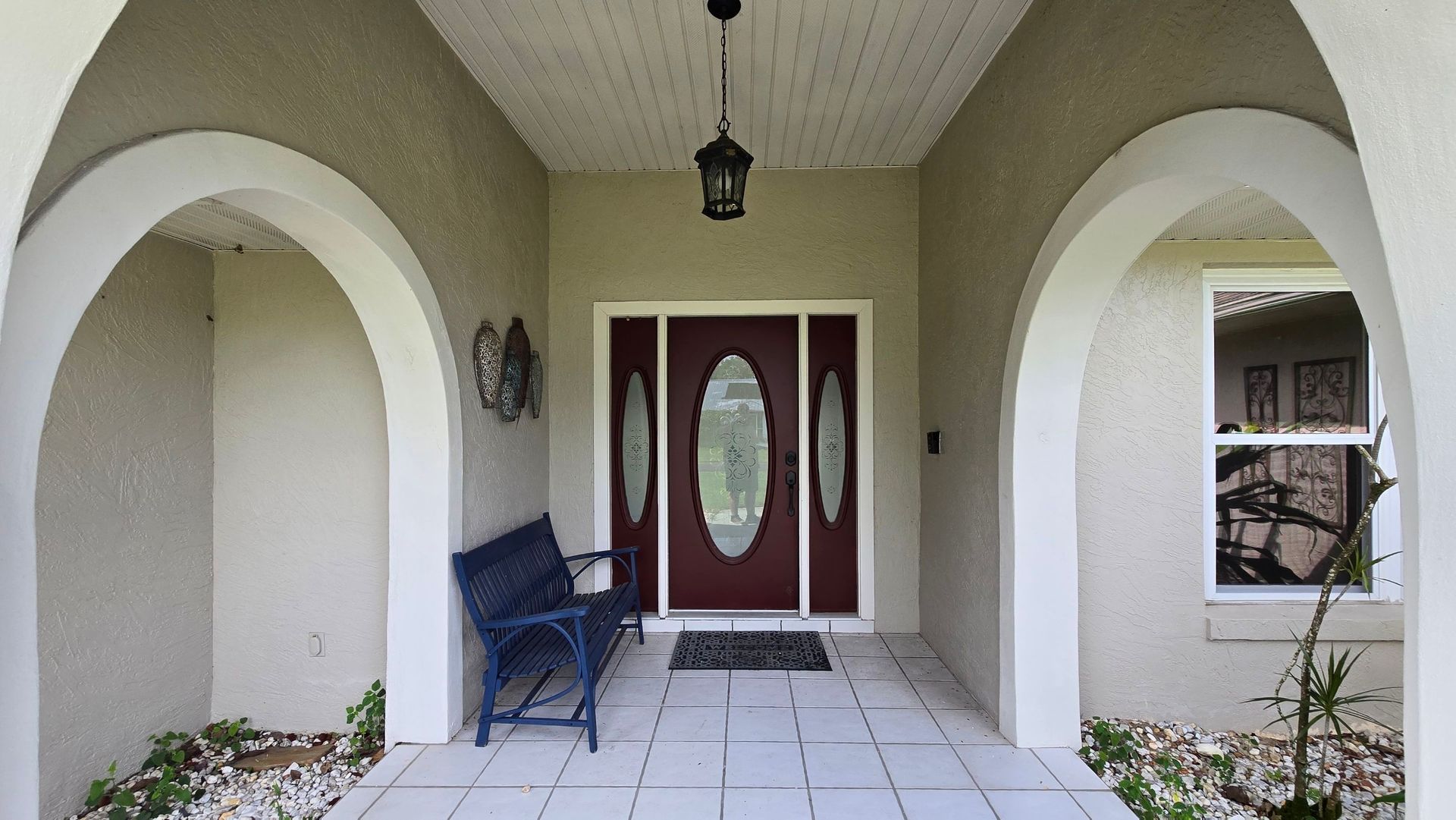 Covered front porch with arched entryways, dark red door, bench, and tiled floor.