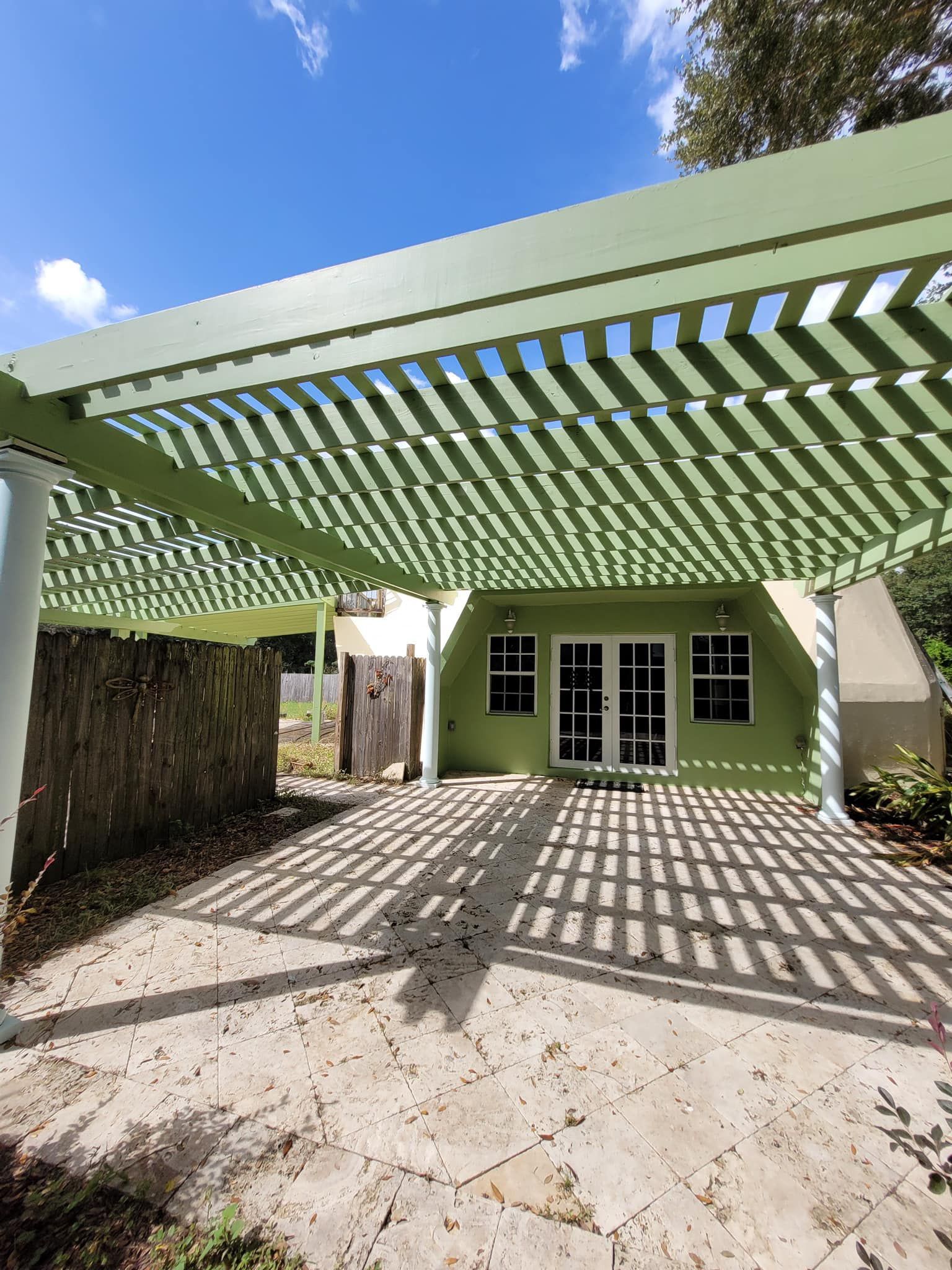 Green pergola casting shadows on a stone patio in front of a light green house with double doors.