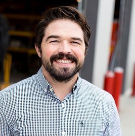Smiling man with dark hair and beard in a blue checkered shirt outdoors