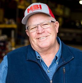 Smiling person in a red-and-gray MBI cap and blue jacket, standing in a workshop.