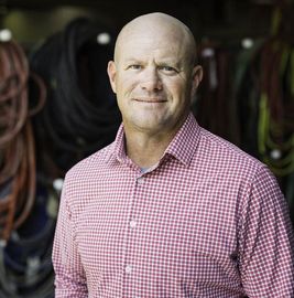 Man in a pink checkered shirt standing in a workshop with coiled hoses in the background