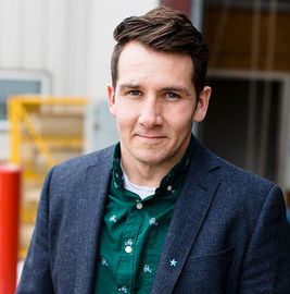 Portrait of a man in a green shirt and blue blazer, standing outdoors with a blurred building behind him