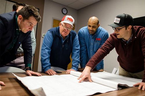 Four workers lean over blueprints on a conference table, discussing a project in an office room.