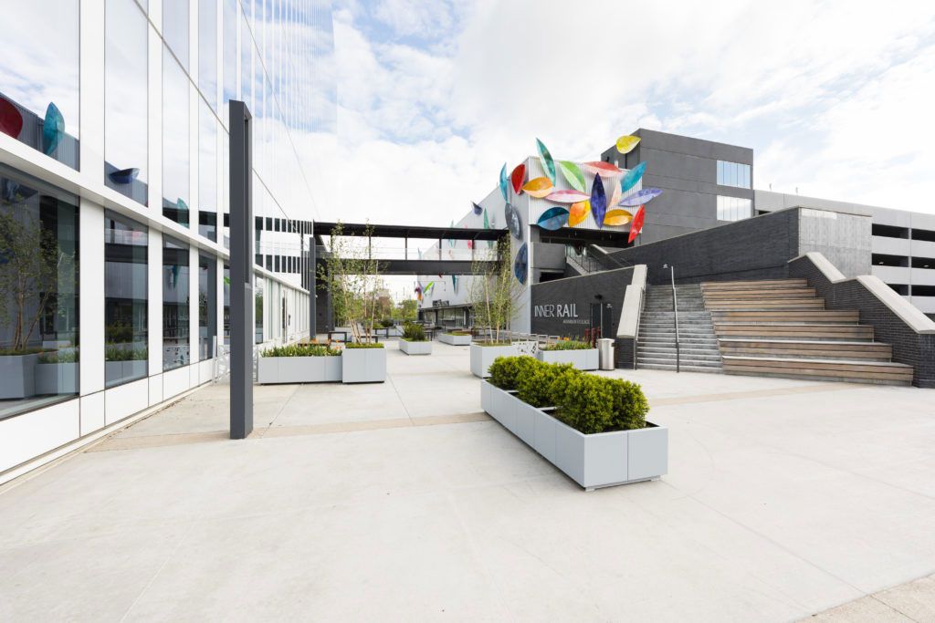 Modern courtyard with colorful sculpture, steps, planters, and glass-front building under a bright sky