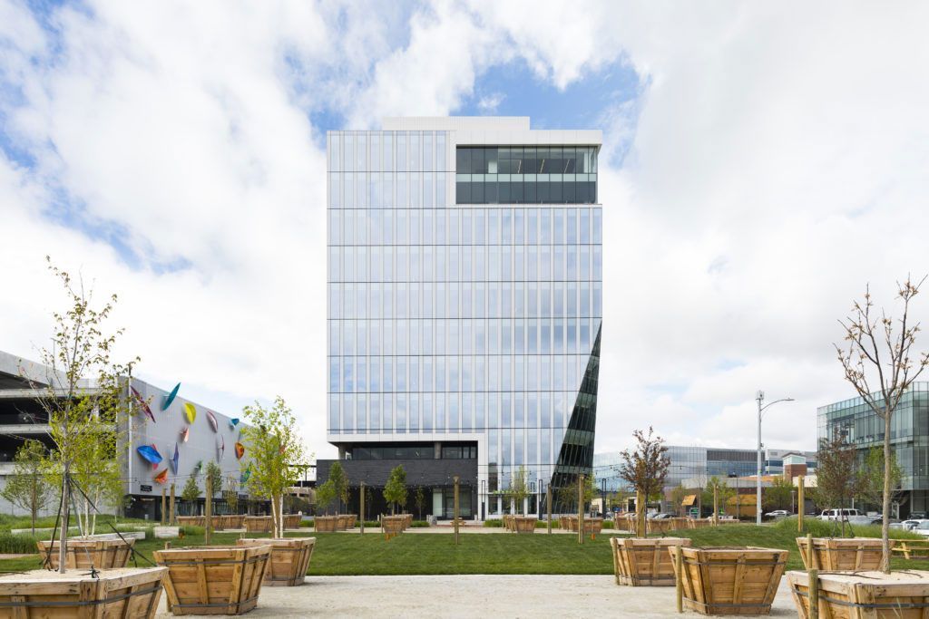 Modern glass office building with landscaped plaza and benches under a bright cloudy sky