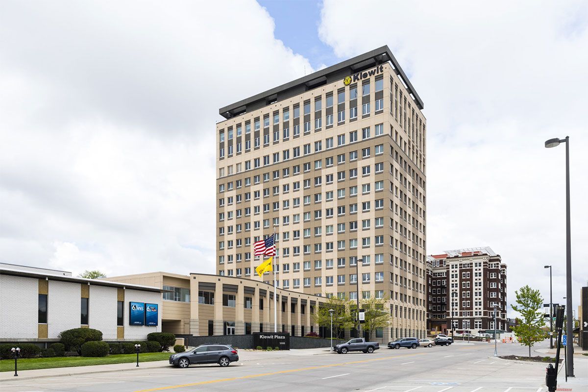 Tall beige office building on a city street under a cloudy sky, with cars parked nearby.