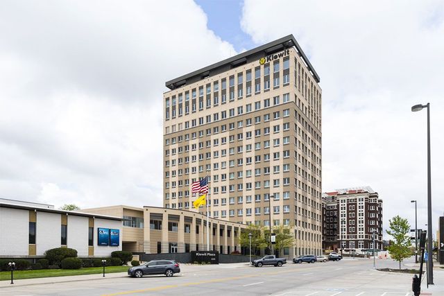 Tall beige office building on a city street under a cloudy sky, with cars and a sidewalk in front