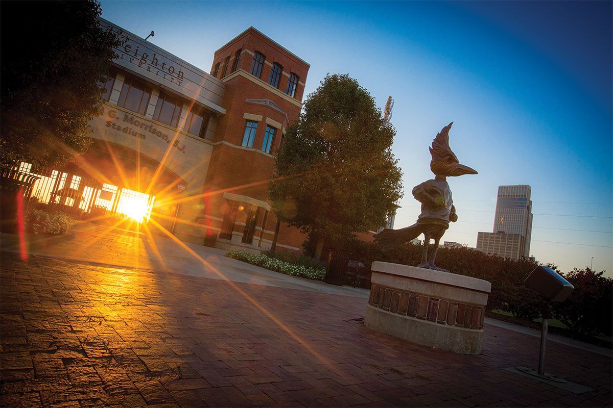 Sunset over a campus courtyard with a brick building, statue, and bright sun flare on the pavement