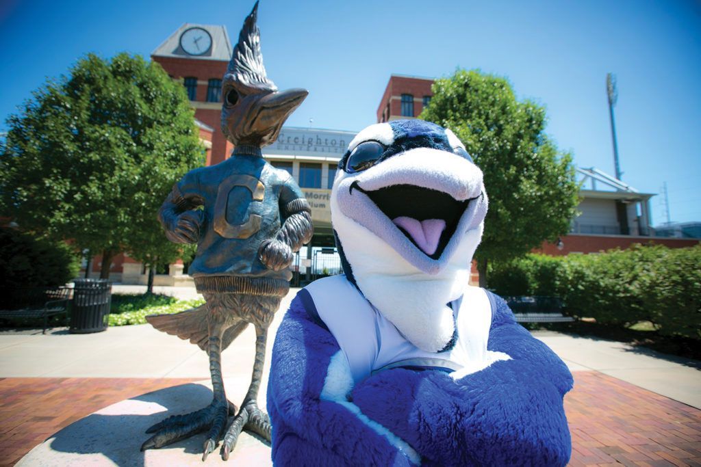 Two bird mascots pose outdoors in front of a building, one blue-and-white and smiling, one gray with a hat.