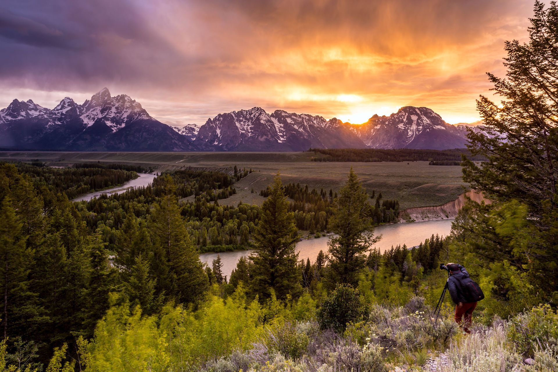 A person is standing on top of a hill overlooking a river and mountains at sunset.