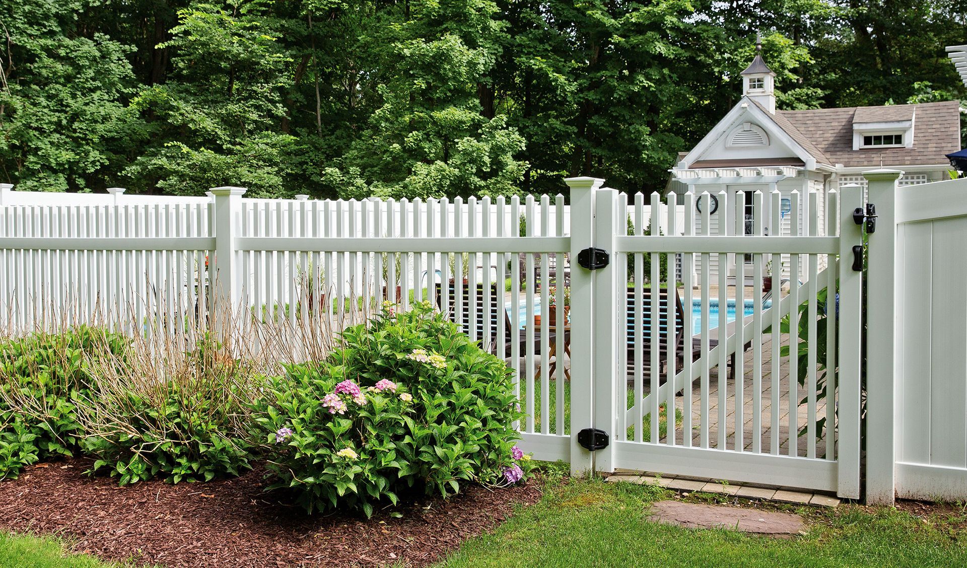 White picket fence and gate beside a landscaped garden with bushes, flowers, and a house in the background