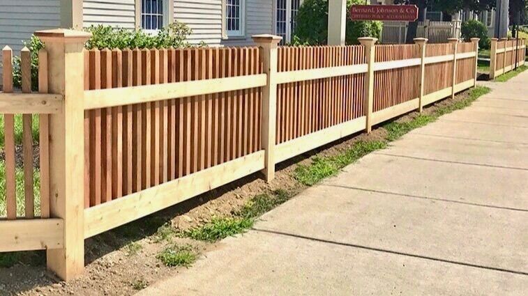 Wooden picket fence along a sidewalk beside a house and green lawn.