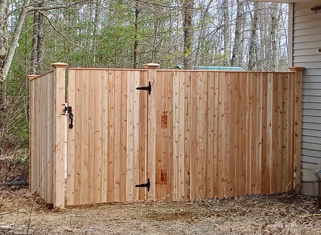 Wooden garden gate and fence beside a house, with trees and leaf-covered ground in the background