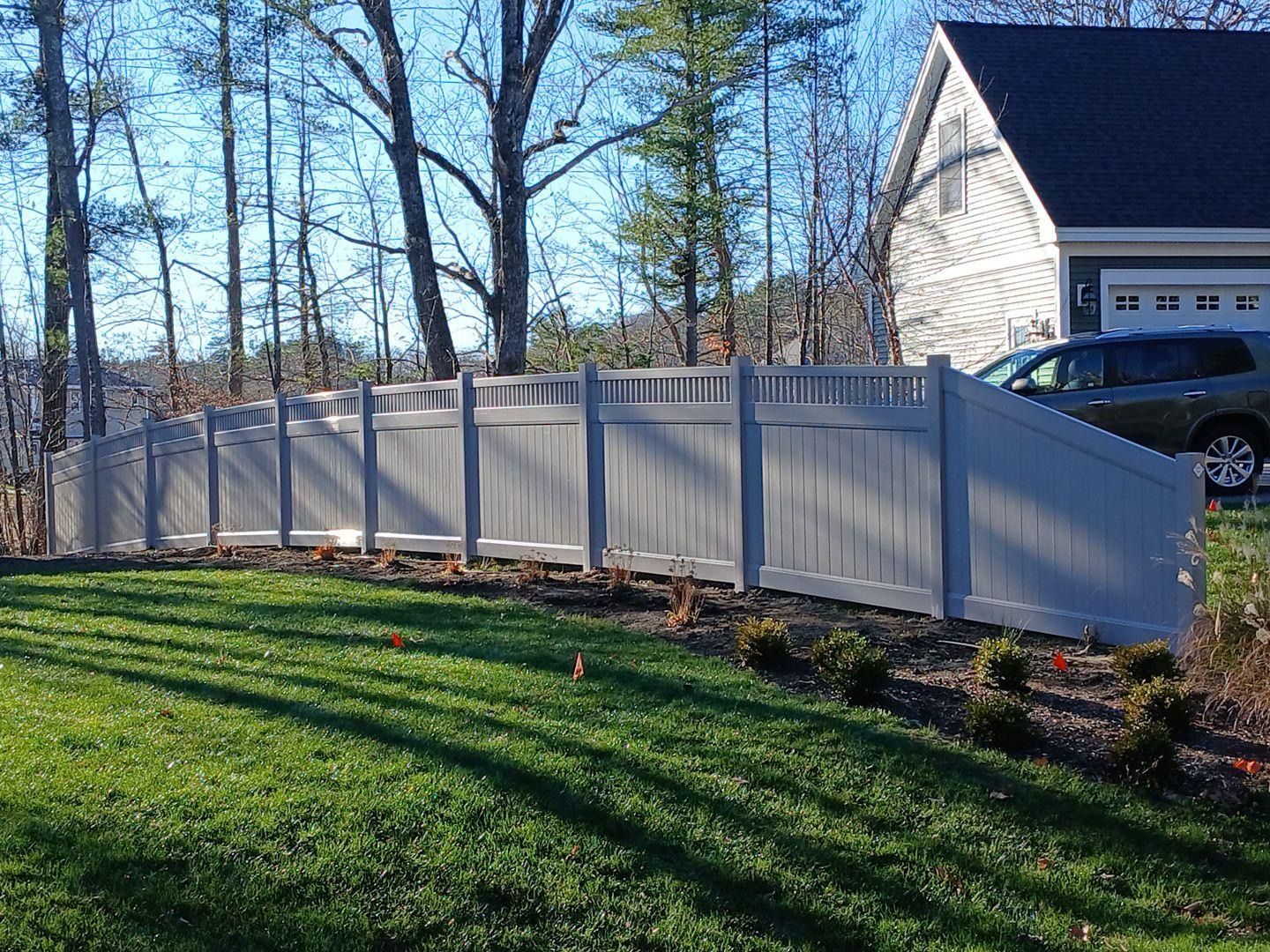 White vinyl fence along a sloped grassy yard beside a house and parked car on a sunny day