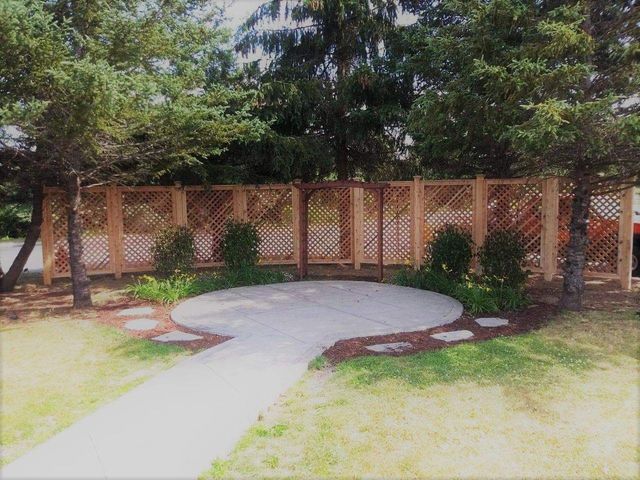 A brick wall with lattice panels behind a curved concrete patio and walkway, surrounded by trees and grass.