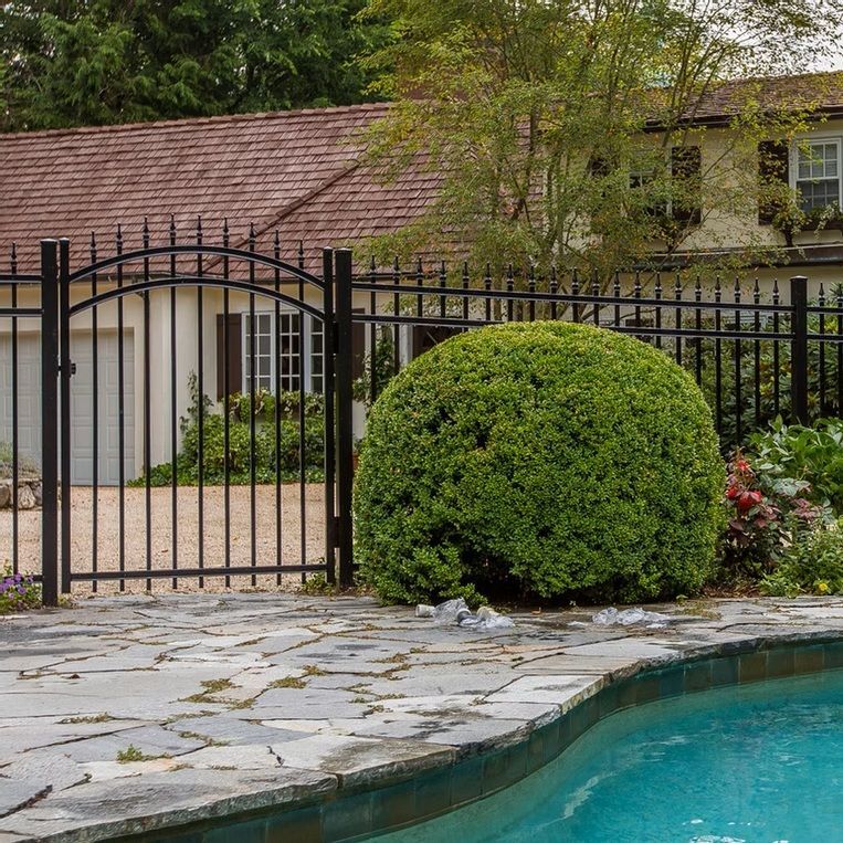 Backyard pool beside stone patio, black metal gate, trimmed shrub, and house in the background