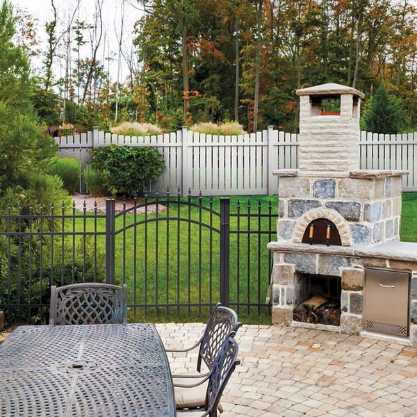 Backyard patio with stone pizza oven, metal table, black gate, white fence, and trees in the background