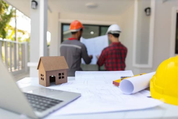Construction workers reviewing plans with a model house, laptop, rolled blueprint, and yellow hard hat on a table.