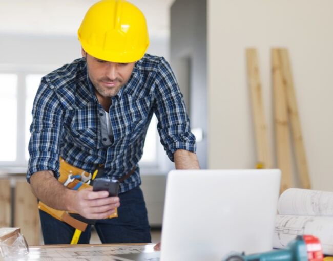 Worker in yellow hard hat leaning over a laptop and checking a phone at a construction site.