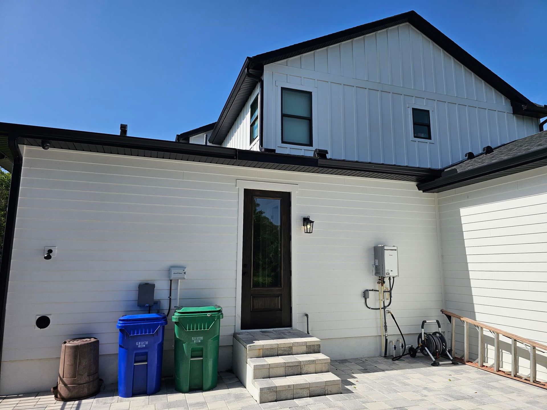 Back patio of a house with trash bins, steps, white siding, and a ladder on the right.