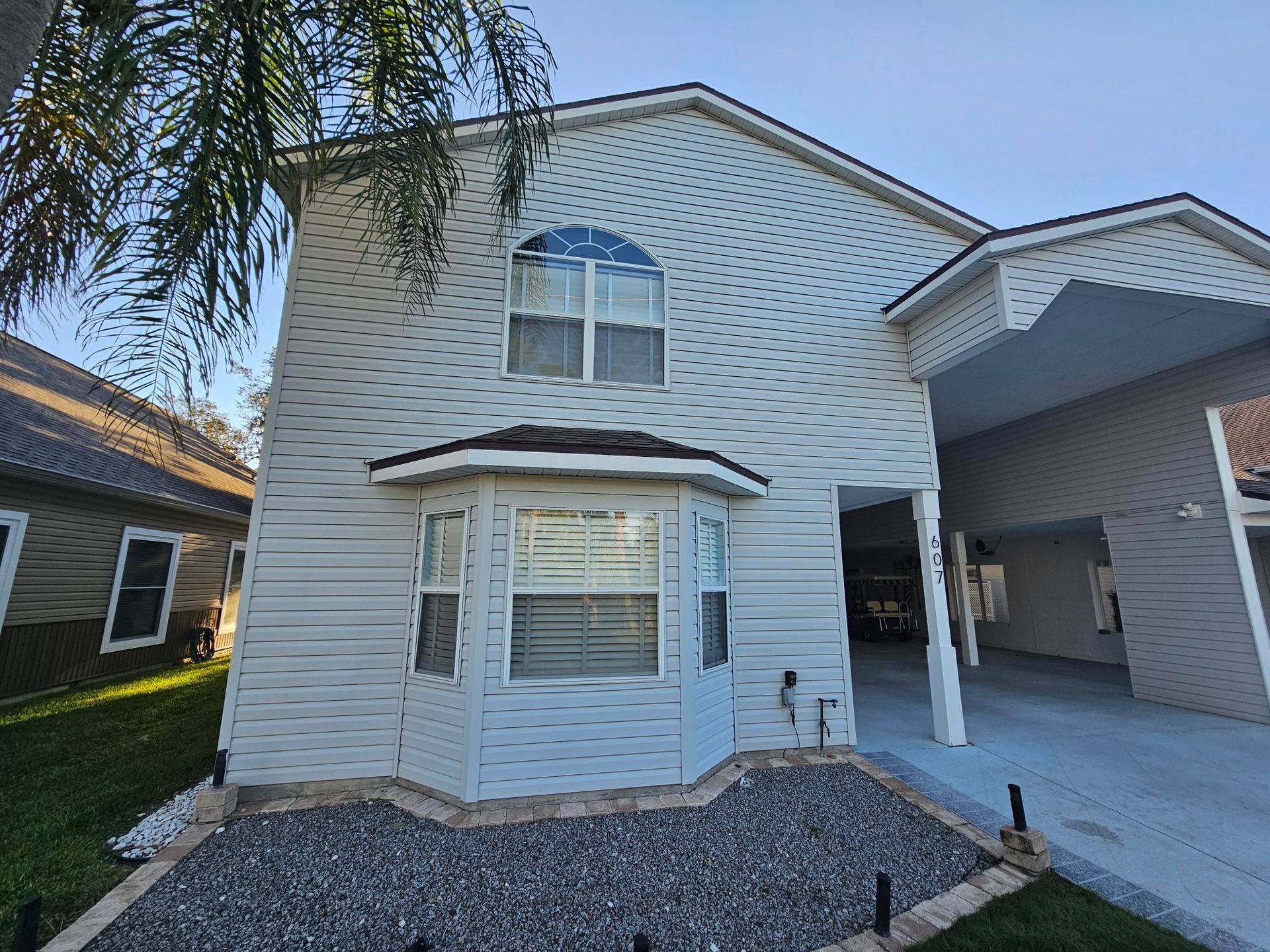 Two-story white house with bay window, gravel front yard, and covered carport on the right.